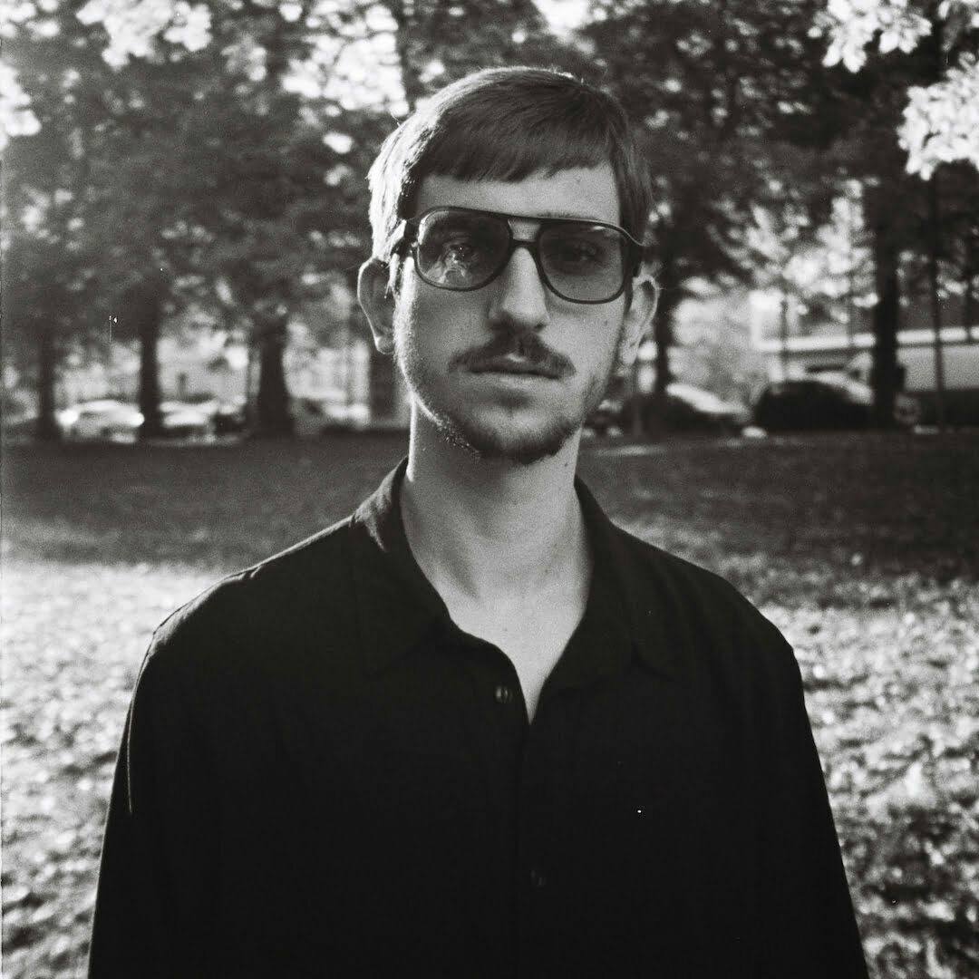 Black and white photo of Madd Rod in a garden wearing sunglasses.